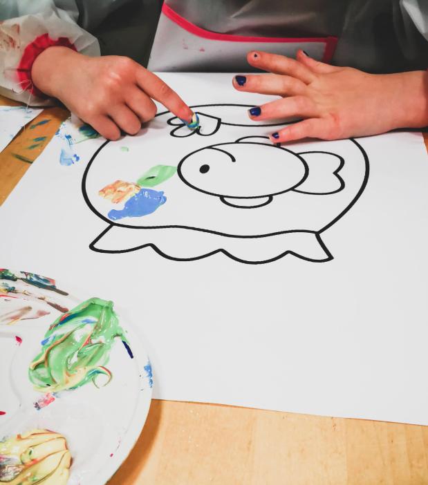 Een kind verft met vingerverf een vis in een bokaal op een blad papier.